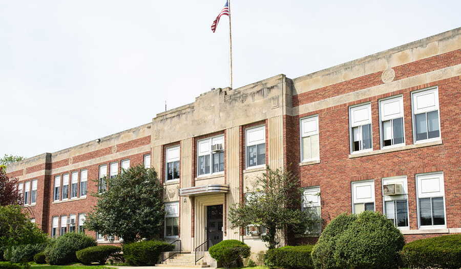 Front view of a two-story red-brick public school building with an American flag on the roof, large windows, and trimmed shrubs and trees along the entrance.