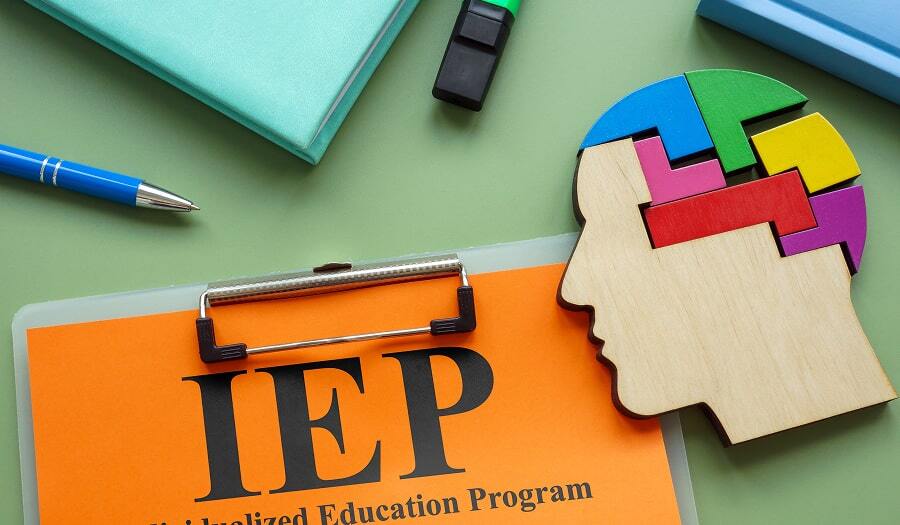Clipboard with a bright orange cover sheet labeled “IEP Individualized Education Program” surrounded by school materials, including a pen, highlighter, notebook, and a wooden puzzle shaped like a child’s head.