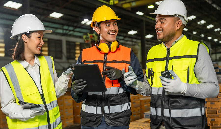 Three warehouse workers in hard hats and high-visibility safety vests stand together on a warehouse floor, smiling as they review information on a tablet.