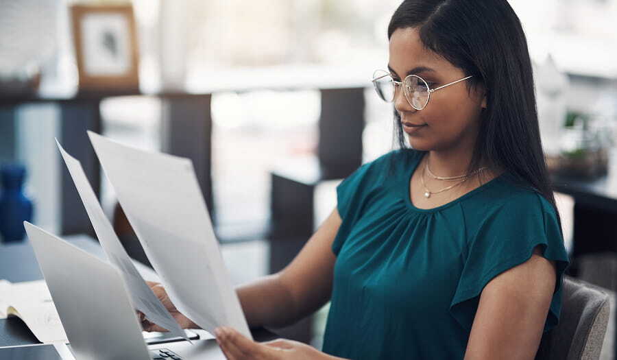 A woman wearing glasses and a teal blouse reviews printed documents at her desk while working on a laptop in a modern office.