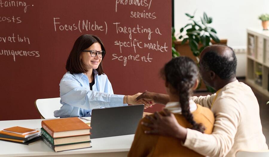 Teacher shaking hands with a parent during a school meeting while their child sits beside them