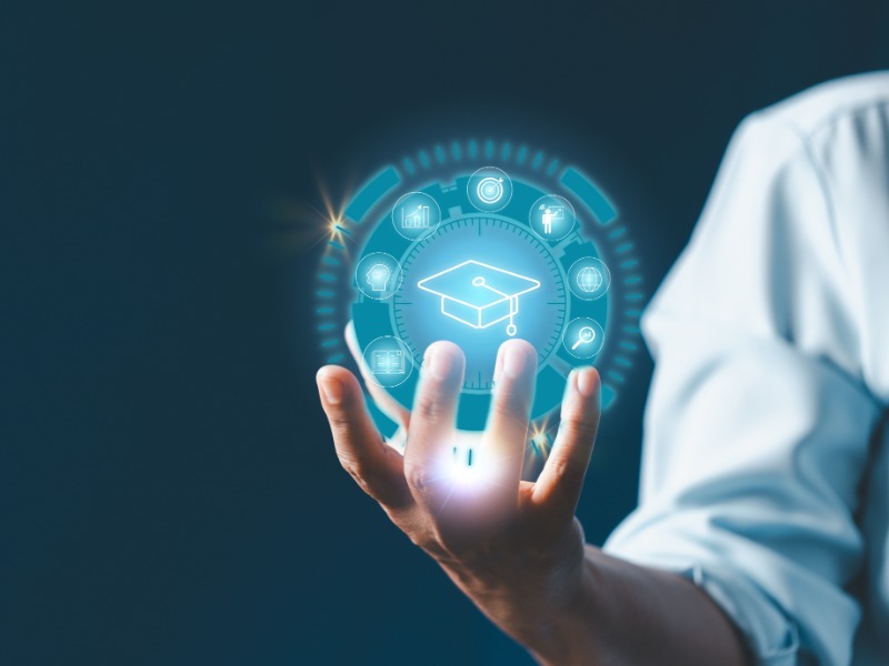 A hand holds a glowing hologram of a graduation cap surrounded by learning icons