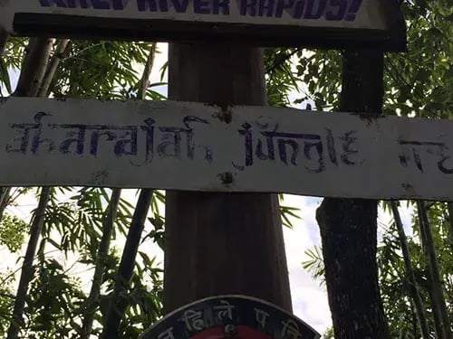 Faded sign with text in English and another script in a forest setting.