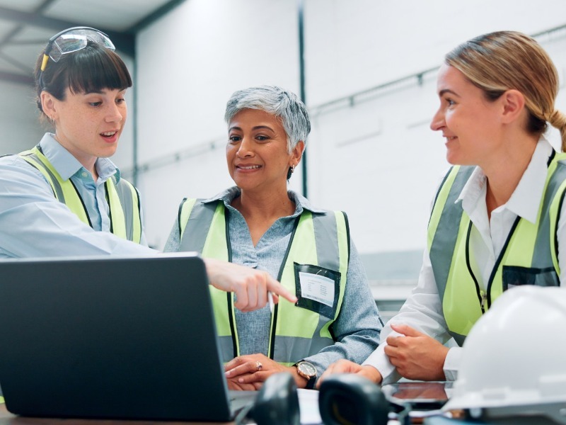 Three women in safety vests review a laptop on a factory floor