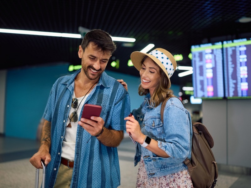 Two travelers checking a smartphone at an airport departures board