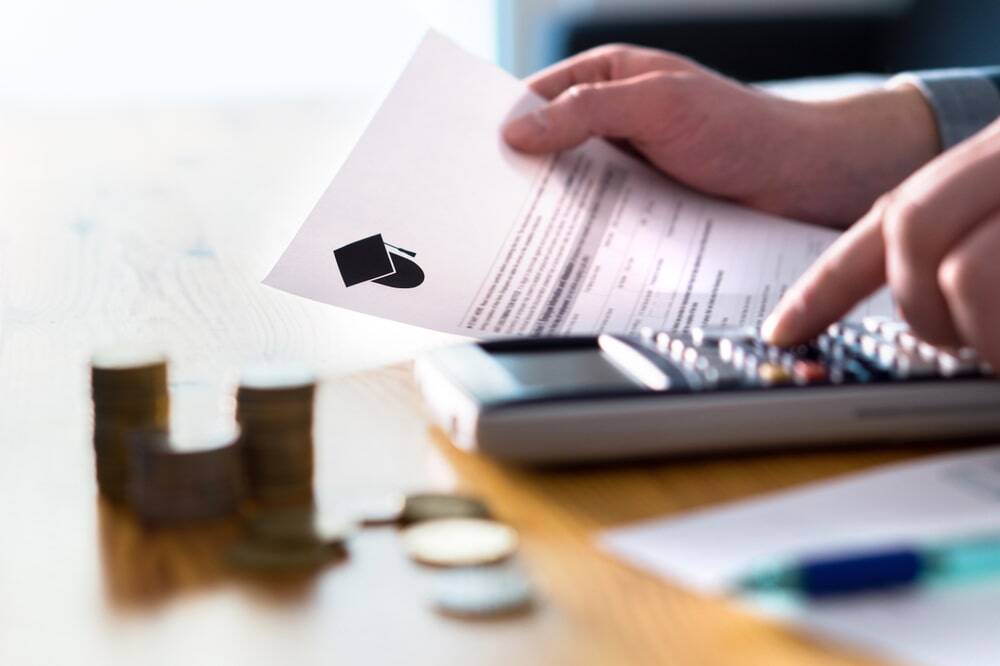 Hand using a calculator beside a school funding document marked with a graduation-cap icon, with stacks of coins on the desk