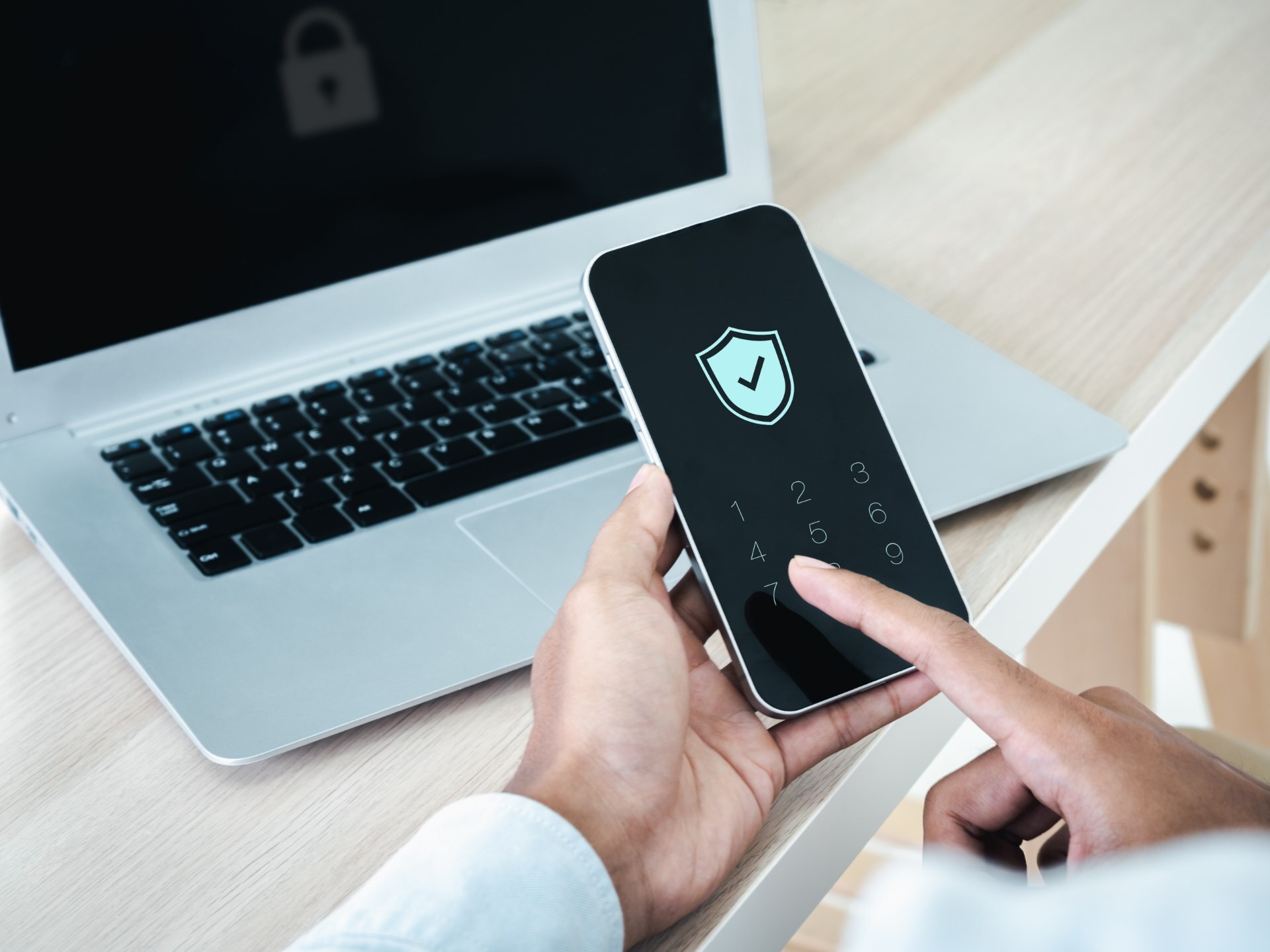 A hand entering a passcode on a phone showing a shield icon, next to a laptop displaying a padlock