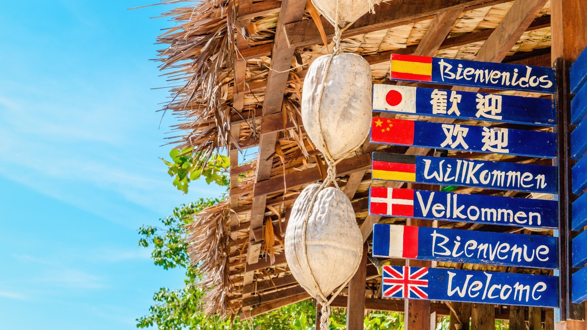 Wooden welcome signs in seven languages, each with a national flag, hanging from a thatched roof