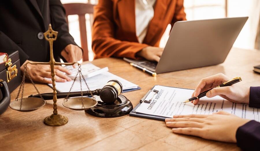 People reviewing and signing legal documents at a table with a gavel, scales of justice, and a laptop.
