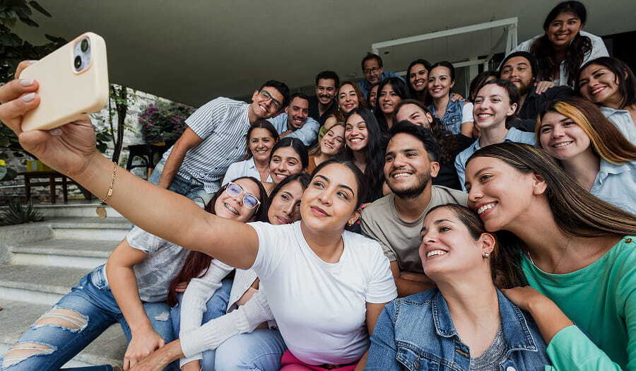 A large group of smiling young Latino adults gathered closely together on outdoor steps while a woman in front takes a selfie with her smartphone.