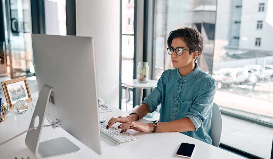Person wearing glasses types at a desktop computer in a bright office, seated at a white desk near large windows.