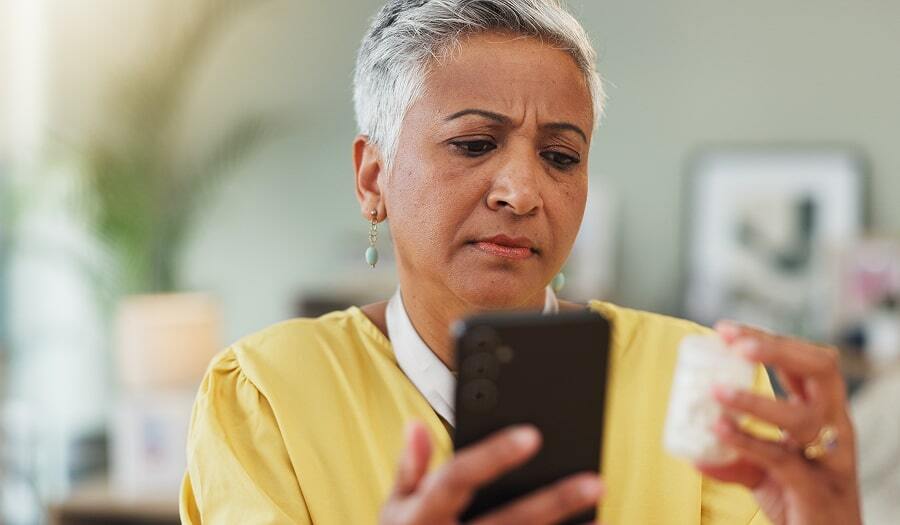 Woman holding a prescription bottle squints at her phone screen, appearing uncertain about her medication instructions