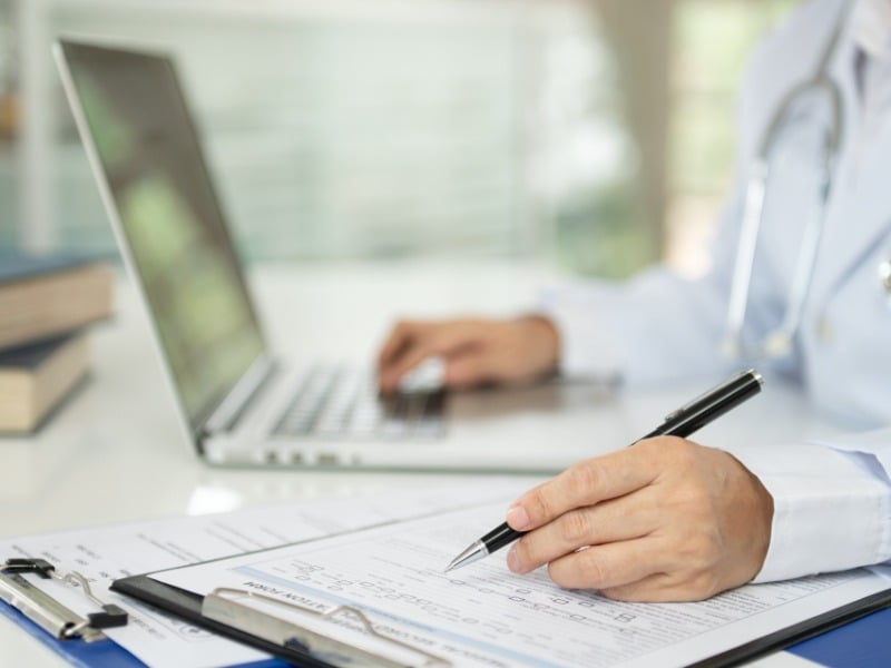 A clinician fills in a medical record form on a clipboard next to a laptop