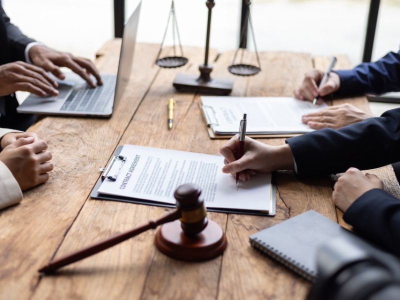 People at a table signing contract agreements, with a gavel and scales of justice
